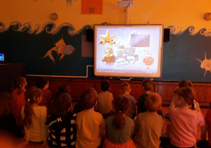 Dzieci z grupy VI i V siedzą na ławeczkach w sali gimnastycznej i oglądają film edukacyjny o Unii Europejskiej.